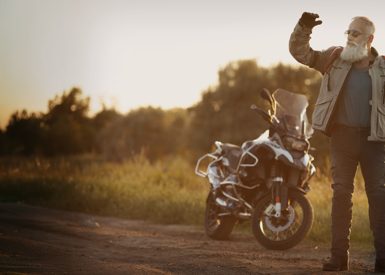 An older rider with a grey beard waving beside his adventure motorcycle at sunset